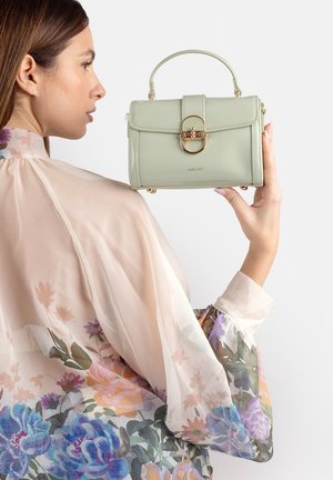 Donna con una camicetta trasparente a fiori che tiene una borsa verde chiaro con chiusura dorata e manico superiore su uno sfondo semplice.