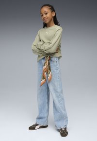 Sudadera verde de manga larga, jeans anchos de color azul claro y una bufanda a rayas naranja y verde atada a la cintura. Zapatillas oscuras con acentos blancos.