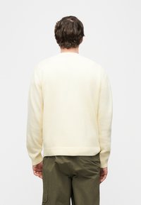 Homme de dos portant un pull en maille crème et un pantalon vert olive avec une poche latérale, debout devant un fond clair uni.