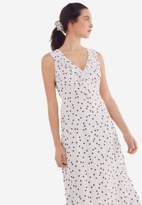 Witte jurk met zwarte stippen, gerimpelde halslijn, mouwloos ontwerp en een vloeiende silhouet. Inclusief een bijpassende haarscrunchie met stippen.