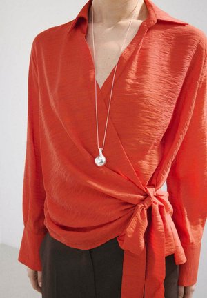 Person wearing a textured long-sleeve orange wrap top tied at the waist, paired with dark pants and a silver teardrop pendant necklace.