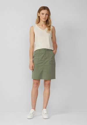 Grüner Baumwollrock mit geradem Schnitt, zwei Fronttaschen, kombiniert mit einem cremefarbenen ärmellosen Oberteil und weißen Sneakers. Glatte Textur.