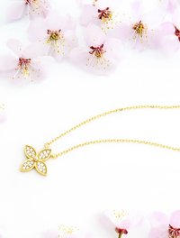 Collier en or avec un pendentif en forme de papillon orné de pierres précieuses transparentes. La chaîne est délicate et texturée, se détachant sur un fond de fleurs roses douces.