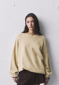 Pulover oversized bej cu modele, cu guler rotund, umeri căzuți și manșete căptușite. Asortat cu o fustă maro în straturi.