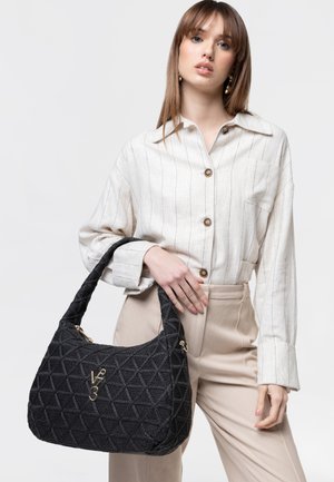Donna con pantaloni beige e camicia bianca a righe sottili che tiene una borsa nera testurizzata con il logo V3 dorato davanti.
