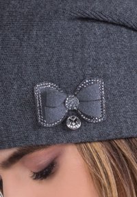 Gray knitted beanie with a decorative bow outlined in rhinestones and a small round charm, worn by a person with light brown hair.