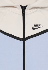 Balta ir šviesiai mėlyna Nike džemperis su juodu užtrauktuku ir juodu Nike logotipu viršutiniame dešiniajame krūtinės plote.