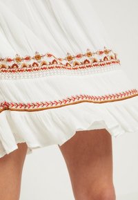 Valkoinen laskostettu hame, jossa on monimutkainen punaisen ja kullan värinen kukka-alkemisto helman kohdalla, ja jossa on röyhelöreunus lisäämässä tekstuuria ja yksityiskohtia.