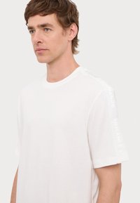 Bílý tričko s krátkým rukávem a kulatým výstřihem, s texturovaným brandingem ARMANI EXCHANGE podél ramene. Měkká bavlněná tkanina s hladkým povrchem.