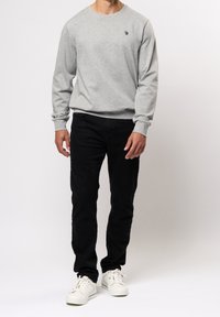 Grå sweatshirt med långa ärmar och rund halsringning, tillsammans med svarta byxor och vita sneakers. Har en liten logotyp på bröstet.