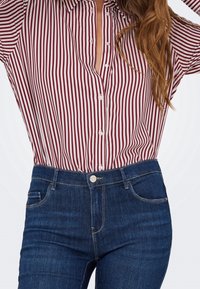 Femme portant une chemise à rayures verticales rouges et blanches boutonnée, rentrée dans un jean bleu foncé taille haute, cheveux partiellement visibles sur l'épaule.