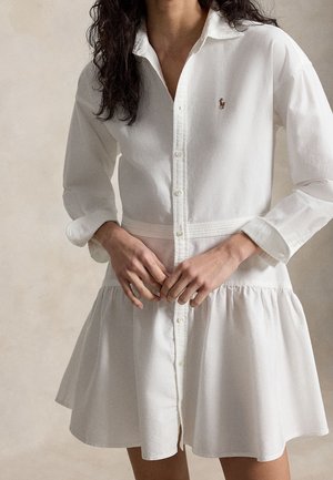 Mujer con cabello largo y oscuro lleva un vestido blanco de manga larga con botones, falda acampanada y un pequeño logo bordado en el pecho.
