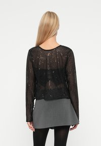 Top preto com lantejoulas de mangas longas, com um design cropped, textura transparente e decote redondo. Combinado com uma saia cinza.