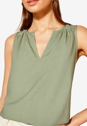 Kvinde iført ærmeløs olivengrøn V-hals top med subtile rynker nær skuldrene og beige bukser, foran en ensfarvet baggrund.