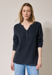 Camicia a maniche lunghe blu scuro con motivo testurizzato, scollatura a V e tre bottoni sul davanti, abbinata a jeans azzurri chiari.