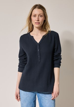 Donker marineblauw langemouwen shirt met een geaccentueerd patroon, V-hals en drie knopen aan de voorkant, gecombineerd met lichtblauwe jeans.