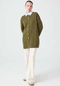 Cardigan a coste verde oliva con una camicia bianca con colletto sottostante, caratterizzato da grandi bottoni e una linea dritta, abbinato a pantaloni a zampa d'elefante color crema.