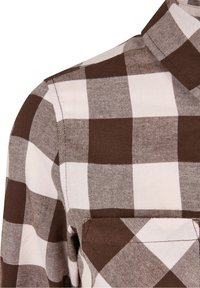 Gros plan sur un tissu de chemise à carreaux marron et blanc montrant les détails du col, de l'épaule et de la poche poitrine.