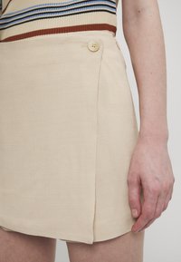 Beige minikjol med texturerad tyg, med en sidoknappdetalj och en asymmetrisk nederkant. Slät yta, avslappnad design.