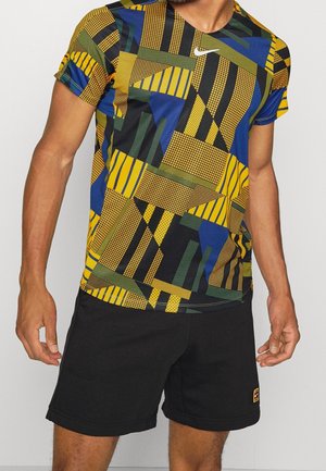 Mann trägt ein mehrfarbiges geometrisch gemustertes kurzärmeliges Hemd und schlichte schwarze Shorts, steht mit entspannten Händen.