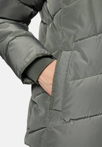 Person's hand inside the pocket of a gray quilted puffer jacket with ribbed cuff visible on the sleeve.