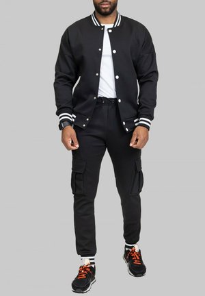 Veste bomber noire avec des poignets et un col à rayures blanches, associée à un jogging cargo noir avec des poches latérales, portée avec des baskets noires.