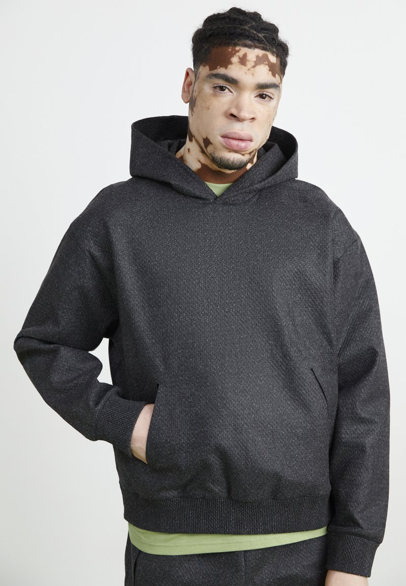 Sudadera con capucha en tela negra texturizada con un bolsillo canguro. Presenta puños y dobladillo acanalados, llevada sobre una capa base color verde claro.