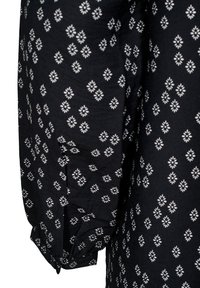 Blouse en tissu noir avec un motif floral blanc répété, manches longues et détails de poignets boutonnés. Texture douce avec un aspect légèrement froissé.