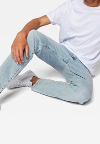 Jeans de mezclilla azul claro con un corte ajustado, costuras visibles y un acabado desgastado, combinados con una camiseta blanca sin estampado y zapatillas blancas.