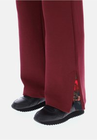 Burgundské tepláky se bočním rozparkem, za kterým se ukrývá květinový vzor, doplněné černými sportovními botami se strukturovanou podrážkou.