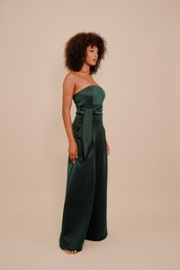 Sieviete ar cirtainiem matiem, ģērbusies tumši zaļā strapless platās kāju kombinezonā ar jostu, stāv sānis uz bēša fona.