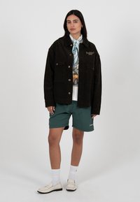 Frau steht und trägt eine schwarze, geknöpfte Jacke, bedrucktes Hemd, türkisfarbene Shorts, weiße Socken, Loafers und einen hellblauen, gemusterten Schal.