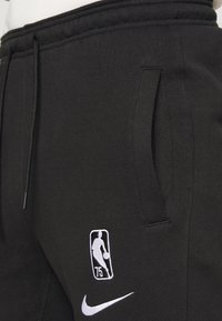 Svarta sweatpants med dragsko i midjan, sidofickor, och broderad NBA 75-årslogga samt Nike swoosh i vitt. Slät tygetextur.
