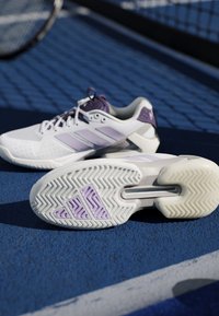 Chaussures de tennis Adidas blanches et violettes sur un court bleu près du filet, une chaussure montre une semelle à motifs avec le logo Adidas visible.