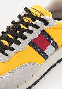 Sneaker gialli con accenti in suede grigio, lacci neri e una striscia logo multicolore sul lato che include rosso, bianco e blu.