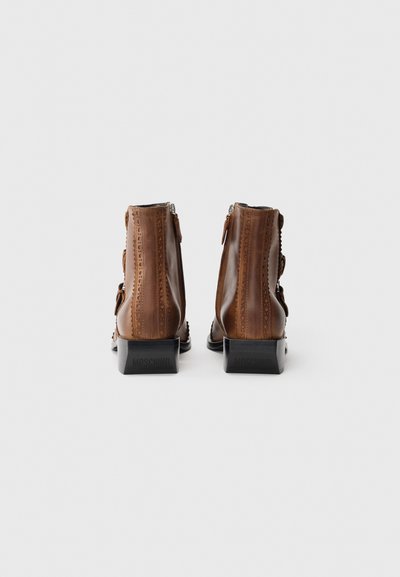 Botines de cuero marrón con un acabado suave, costuras decorativas y un tacón cuadrado negro. Cierre con cremallera en la parte trasera para facilitar su uso.