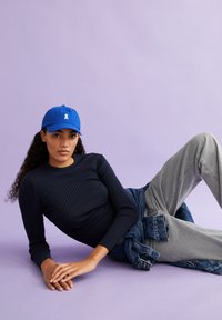 Blaues Baseballcap mit Logo, schwarzes geripptes Langarmoberteil, hellgraue Hose und Jeansjacke, die um die Taille gebunden ist, vor einem lila Hintergrund.