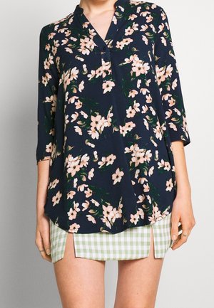 Femme portant un chemisier bleu marine avec imprimé floral rose et manches 3/4 sur une mini-jupe à carreaux verts et blancs avec fentes devant.