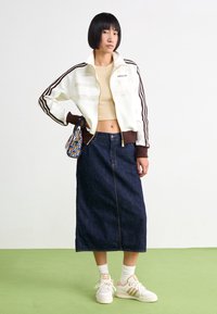 Młoda kobieta w kremowej dżersejowej kurtce Adidas i beżowym topie, ciemnoniebieskiej spódnicy midi, białych sneakersach, trzymająca wzorzysty torebkę na zielonej podłodze.