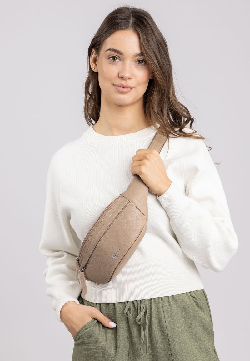 Beige leren schoudertas met een slank, gebogen ontwerp en ritssluiting, gedragen over een witte trui en groene gestructureerde broek.