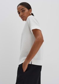 White textured knit top with short sleeves and a loose fit. Features a side slit, worn tucked into dark pants for contrast.