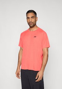 Lyhythihainen urheilupaita kirkkaassa korallin sävyssä, jossa on teksturoitu pinta ja pieni musta logo. Rentouttava istuvuus ja pyöreä kaula-aukko.