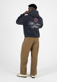 Marineblauwe hoodie met witte tekst en een rood kruisembleem op de achterkant. Gecombineerd met een bruine broek en lichtgekleurde schoenen.