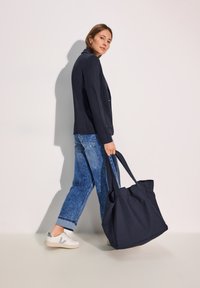 Marinblå axelväska med två långa remmar, tillverkad av slitstarkt tyg. Modellen bär en marinblå jacka och ljusblå jeans med synliga fickdetaljer.