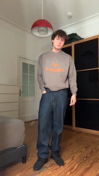Felpa grigia con un logo arancione "LEGACY", abbinata a jeans blu larghi e sneakers nere, ambientata in una stanza con pavimenti in legno.