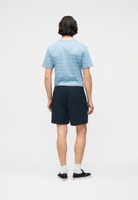 Άνδρας όρθιος με την πλάτη στραμμένη, φοράει μπλε ριγέ μπλουζάκι, ναυτικά σορτς με πίσω τσέπη, λευκές κάλτσες και μαύρα αθλητικά παπούτσια.