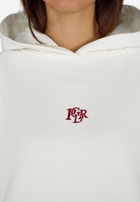 Sudadera con capucha blanca que presenta una tela texturizada y un pequeño logo rojo bordado en el pecho. Diseño sencillo con un corte relajado.