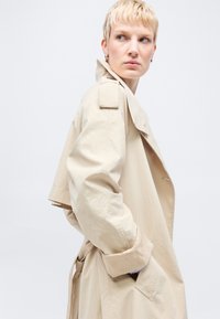 Ξανθό άτομο που φορά υπερμεγέθη μπεζ καμπαρντίνα με ψηλό γιακά, με τα χέρια στις τσέπες, κοιτάζοντας πάνω από τον ώμο μπροστά σε ένα απλό ανοιχτό φόντο.