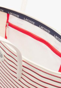 Borsa a tracolla a righe bianca con strisce rosse e nere. Presenta una tasca rossa con zip, fodera in rete e una tracolla blu con logo.