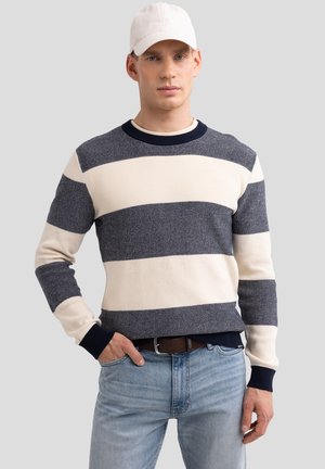 Gestreifter Pullover in Marineblau und Creme, Rippstruktur, runder Ausschnitt, lange Ärmel, kombiniert mit hellblauen Jeans und einer beigen Mütze.